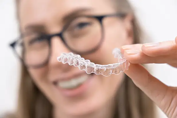 Woman smiling and holding a clear aligner close to the camera, showcasing its transparency and dental alignment benefits.